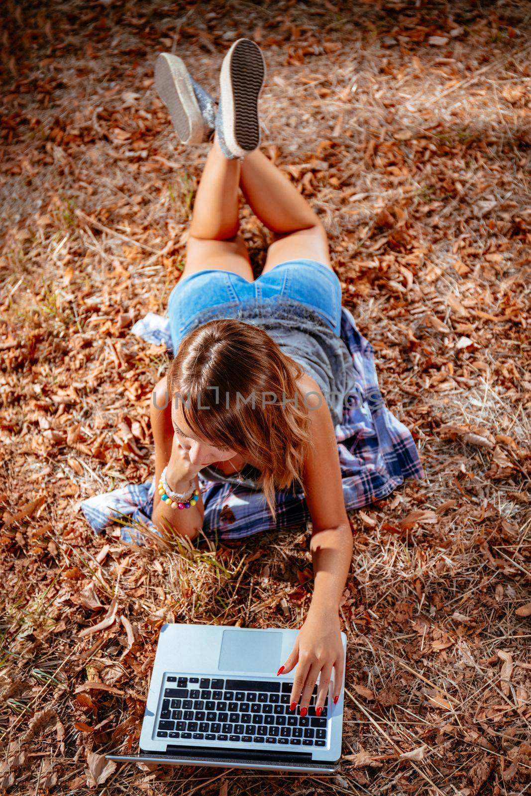 Cute young woman lying down on the withered grass in early autumn sunny day and using laptop.