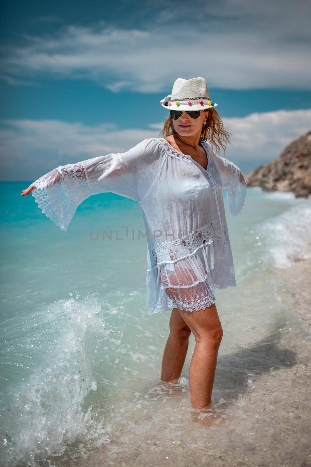 Beautiful young woman in thin wet dress having fun on the beach. She is standing with open arms and looking away with smile on her face. 