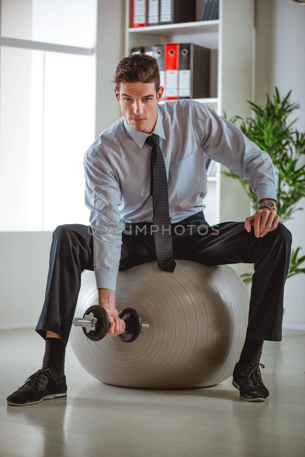 The young businessman sitting in the office on pilates ball and doing exercise with dumbbells.