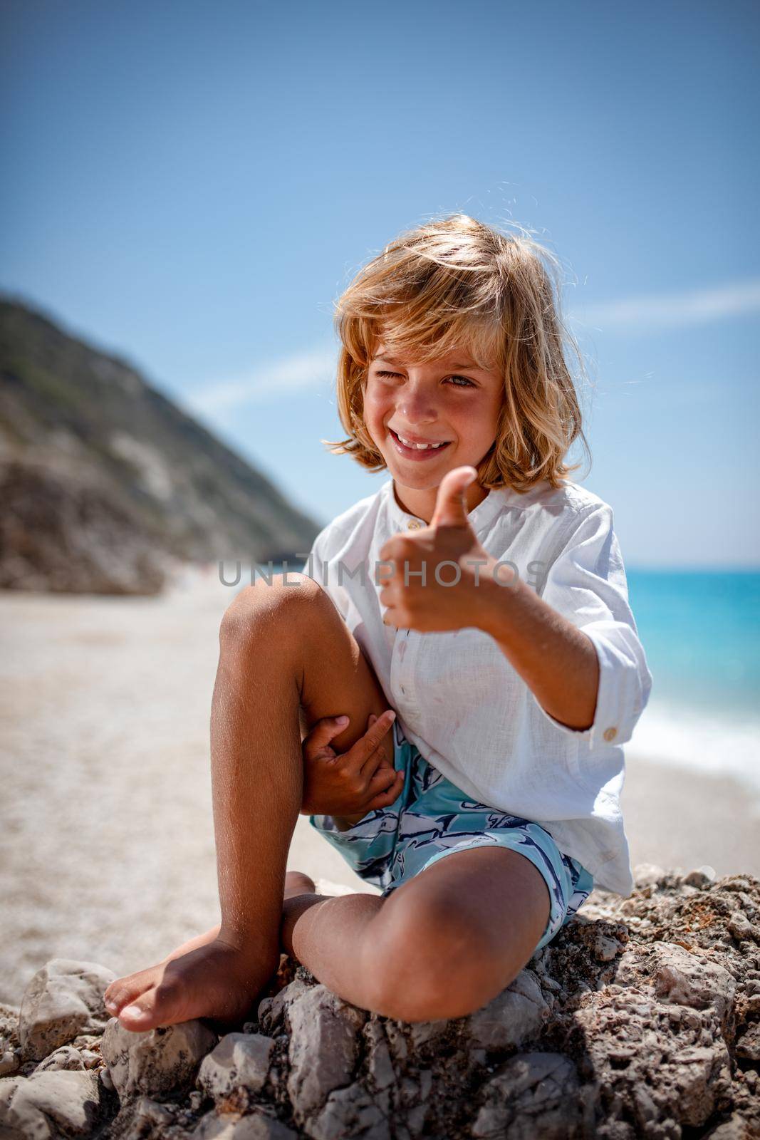 Cute boy is sitting on the sea rock on the beach, showing tumb-up and smiling looking at camera.
