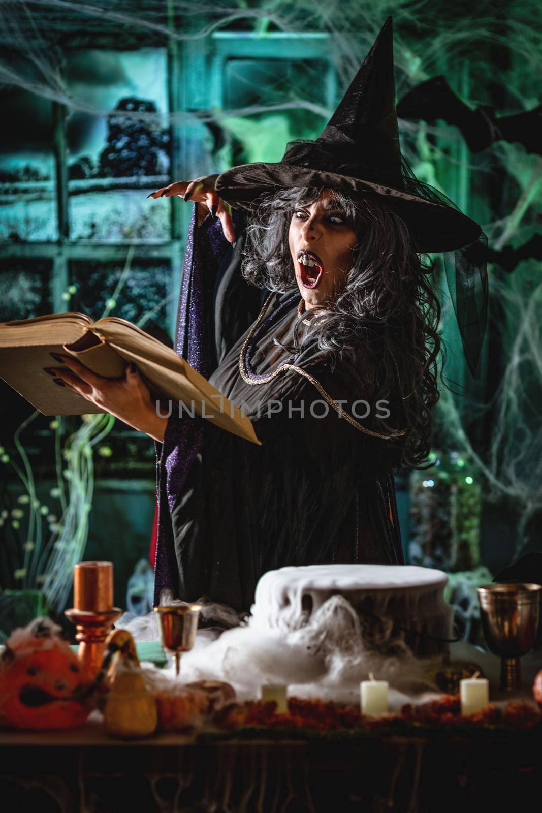 Witch with awfully face in creepy surroundings and smoky green background reading recipe of magic drink sends evil. Halloween concept.