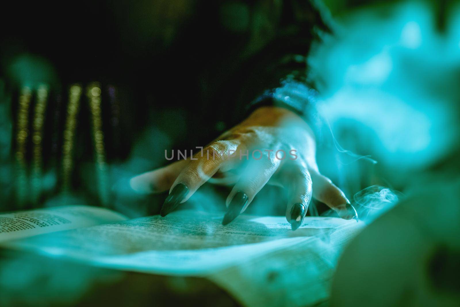 Close-up of a witch's hand with pointed black nails on smoky book of magic recipe and with a magic wand.