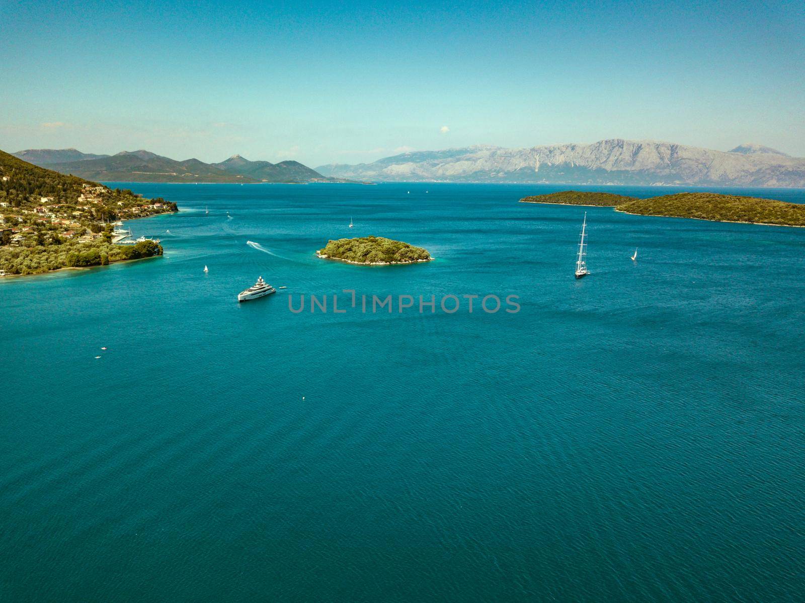 Aerial view on the blue sea with little town on the shore, little island and sailing boats floating. Beautiful natural seascape at the summer time. High angle view on the Nydri bay and a surrounding little islands on the eastern coast of the island of Lefkada, Greece. 