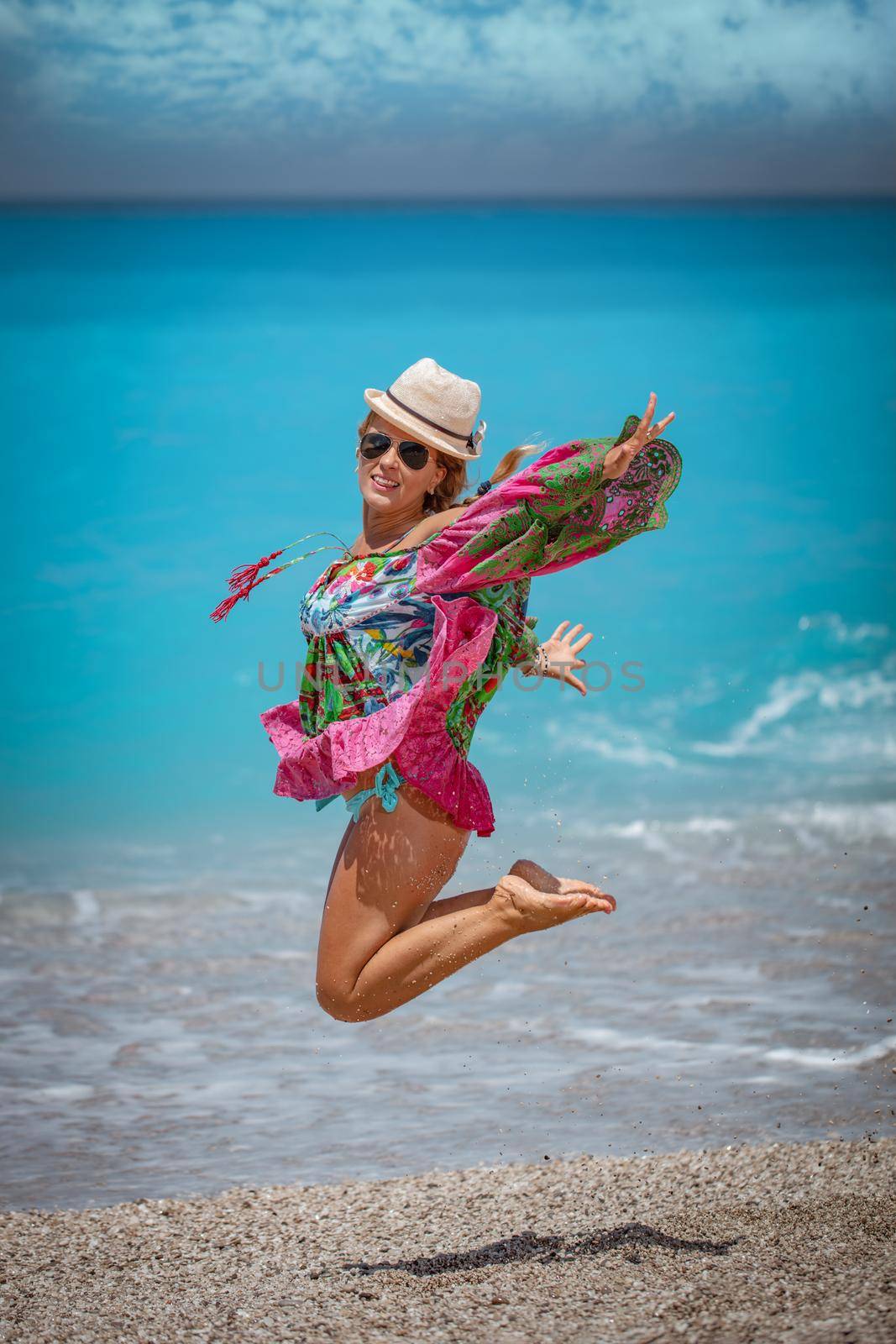 Beautiful young woman in colorful dress is playing in the shore on the beach during the hot summer vacation day. She is enjoying and jumping on the beach. 
