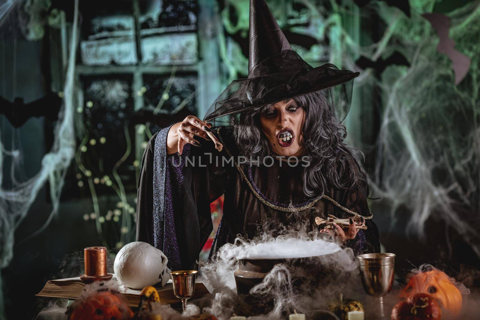Witch with awfully face in creepy surroundings full of cobweb with skull in one and blackbird in the other hand sends evil thoughts. Halloween concept.