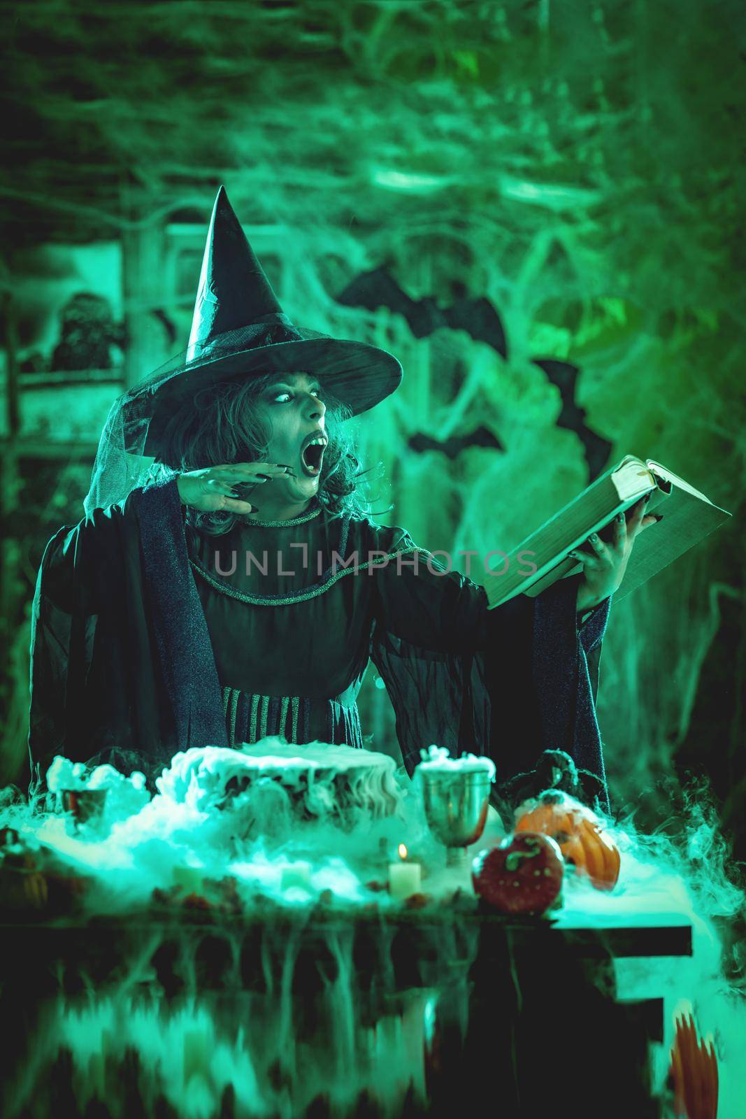 Witch with awfully face in creepy smoky green surroundings full of cobweb is reading recipe of magic drink. Halloween concept.