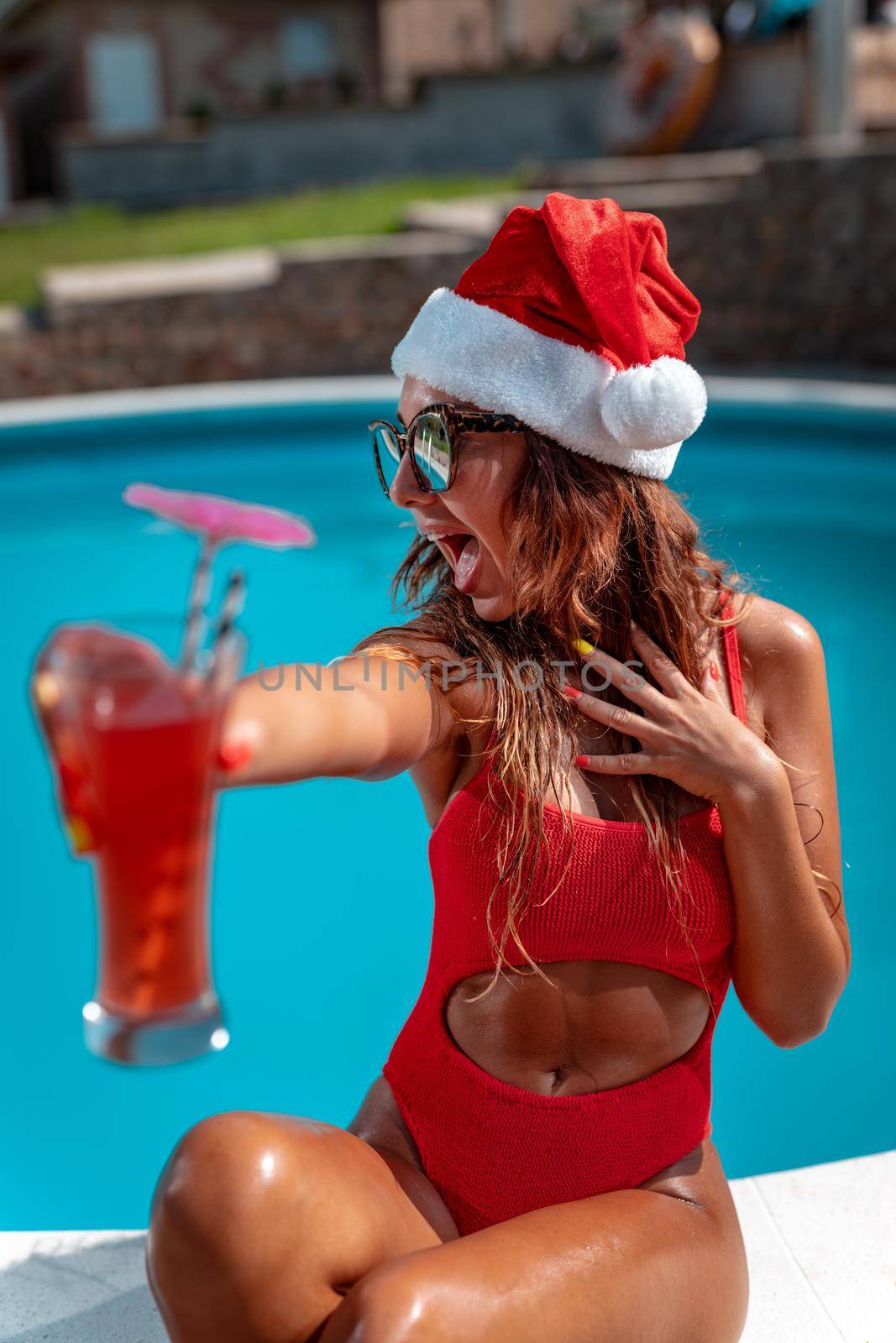 Young beautiful woman near the swimming pool in Santa Claus hat celebrating New Year and Christmas in hot country with glass of cocktail.

