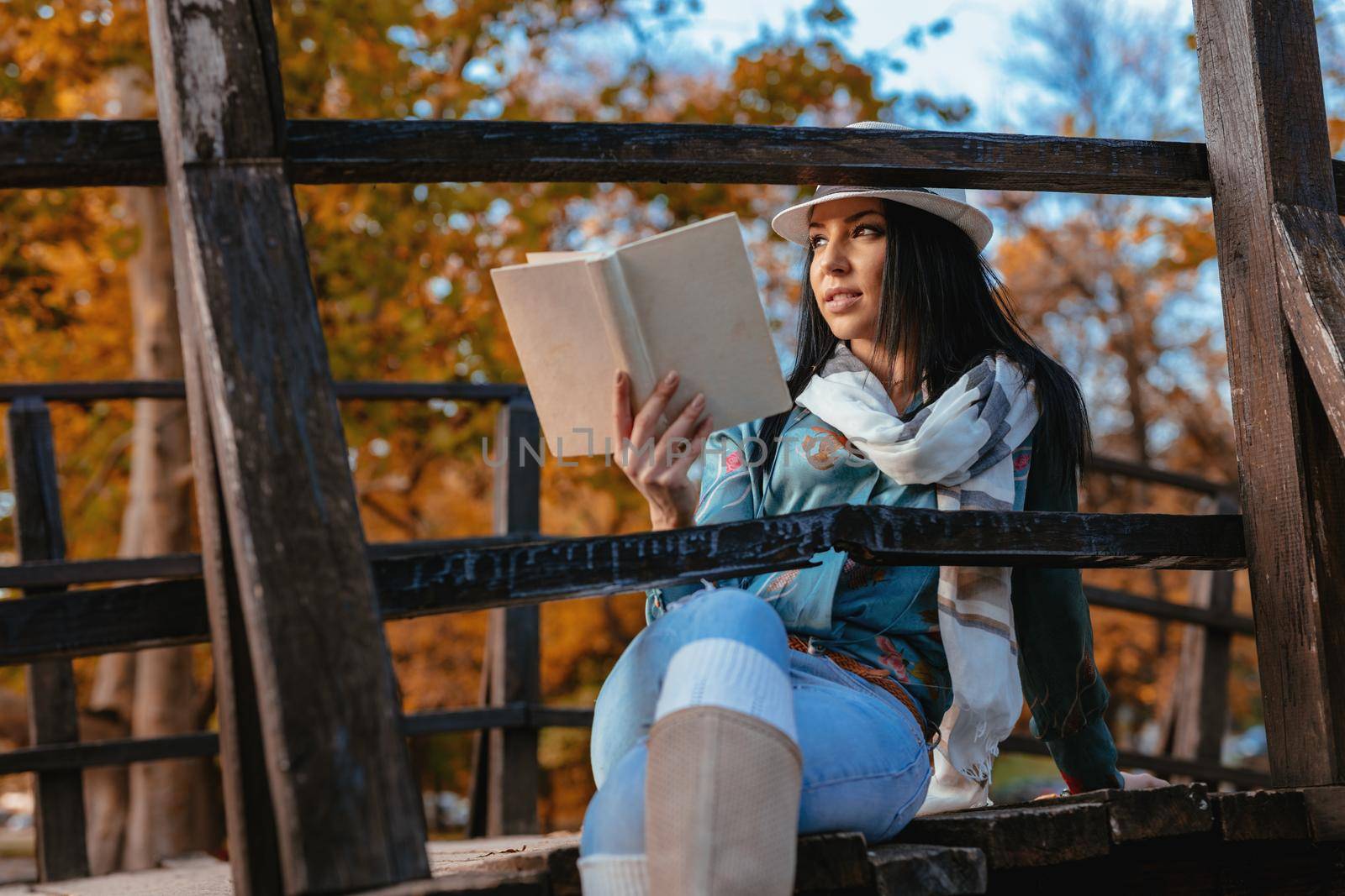 Young happy woman on a wooden bridge over the little lake in the park, enjoying in a beautiful autumn golden day with a book.