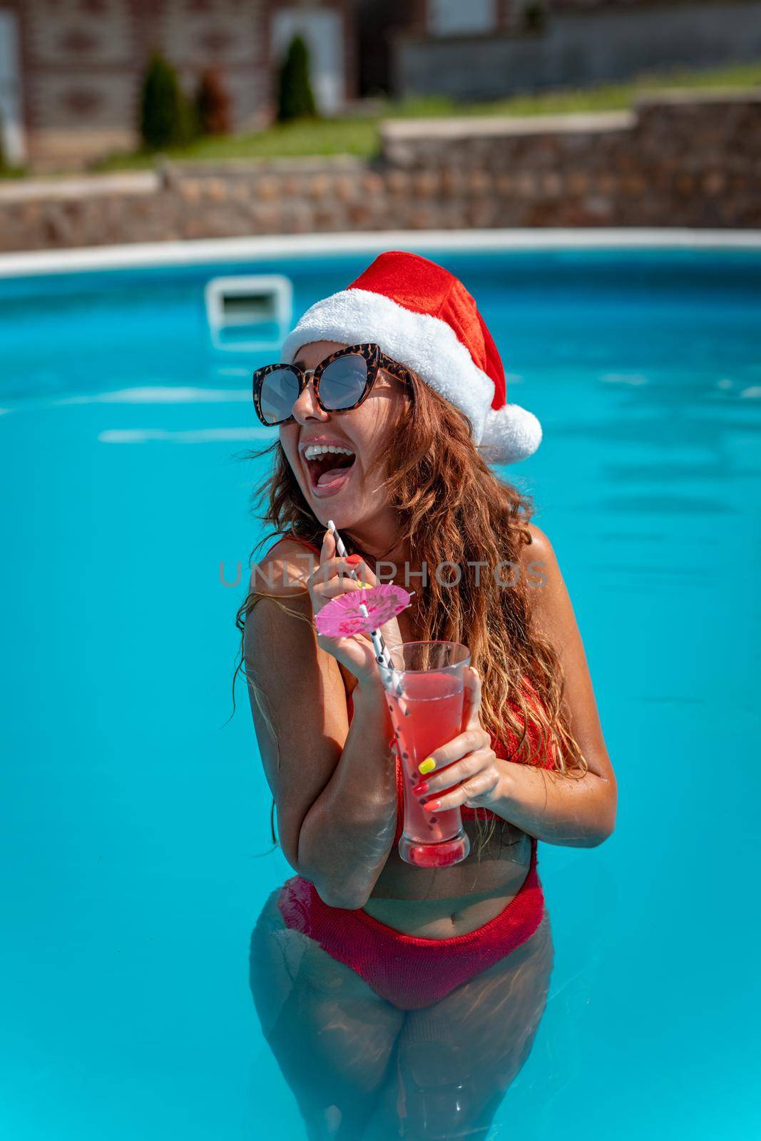 Young beautiful woman near the swimming pool in Santa Claus hat celebrating New Year and Christmas in hot country with glass of cocktail.
