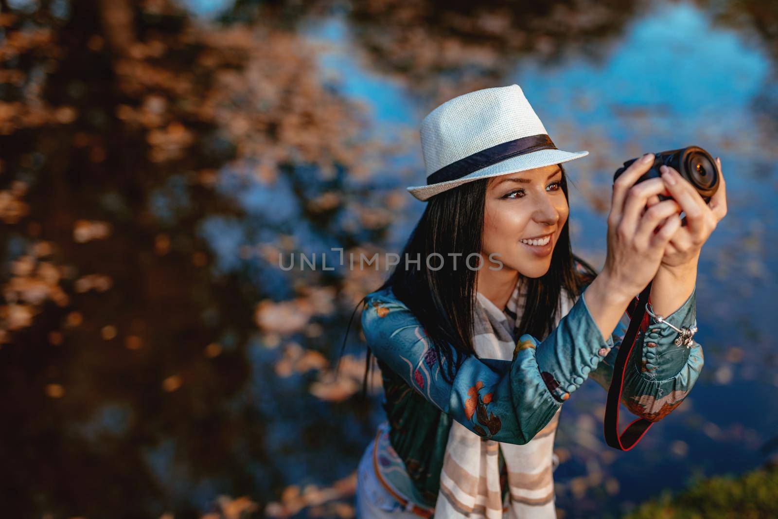 Happy woman photographer holding professional digital camera and taking photo near the lake in autumn park.  