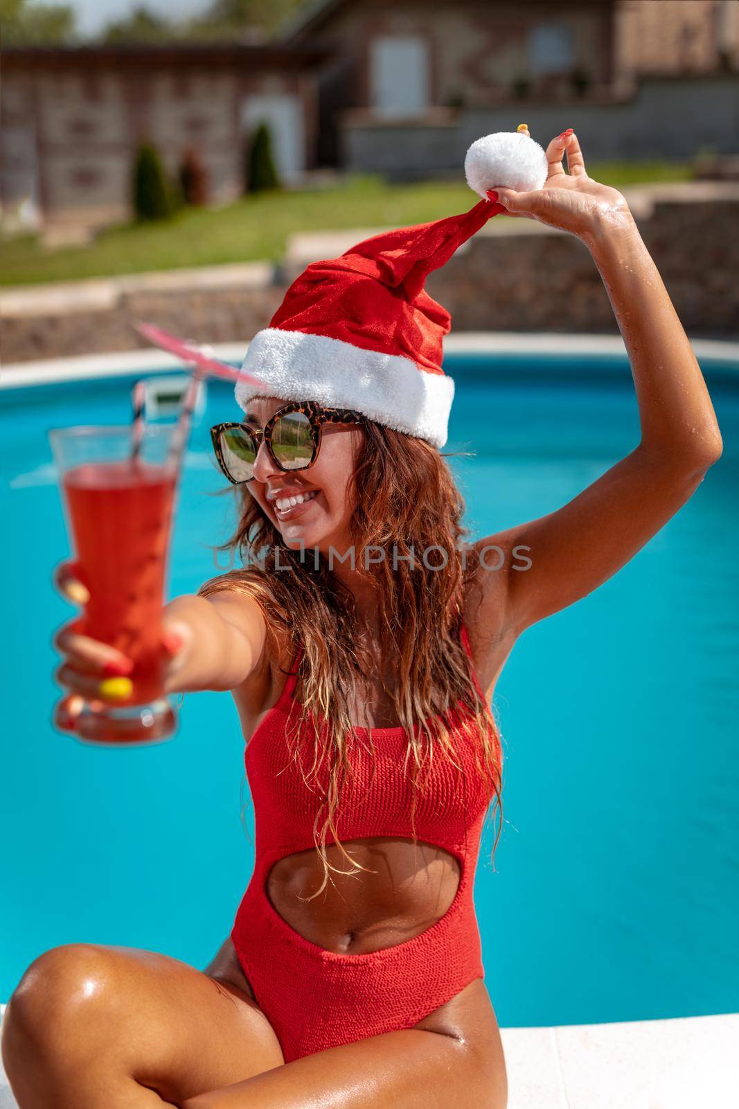 Young beautiful woman near the swimming pool in Santa Claus hat celebrating New Year and Christmas in hot country with glass of cocktail.