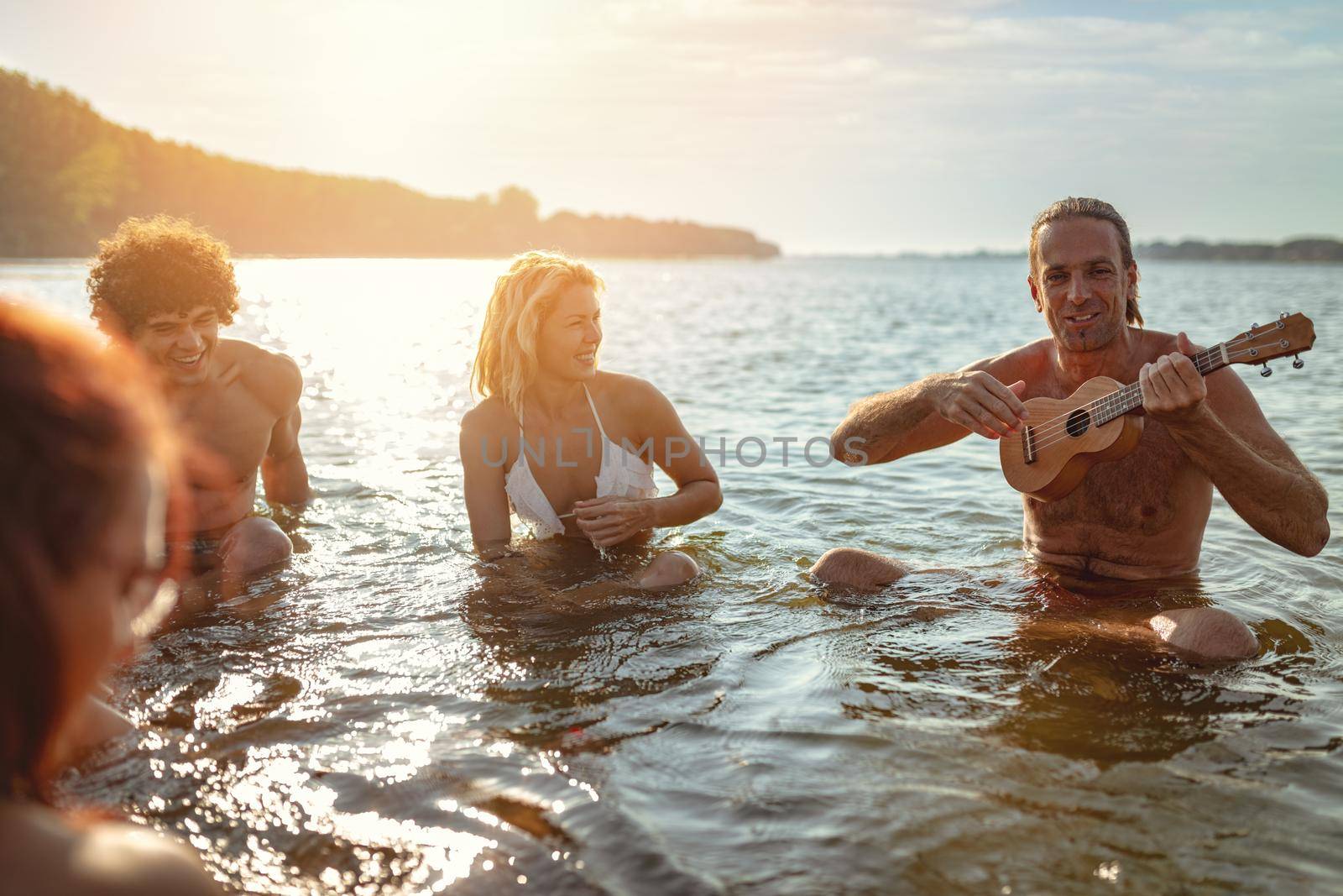 Happy young people having a great time together at the beach. They are sitting in the water and man is playing ukulele and singing. Sunset over water.