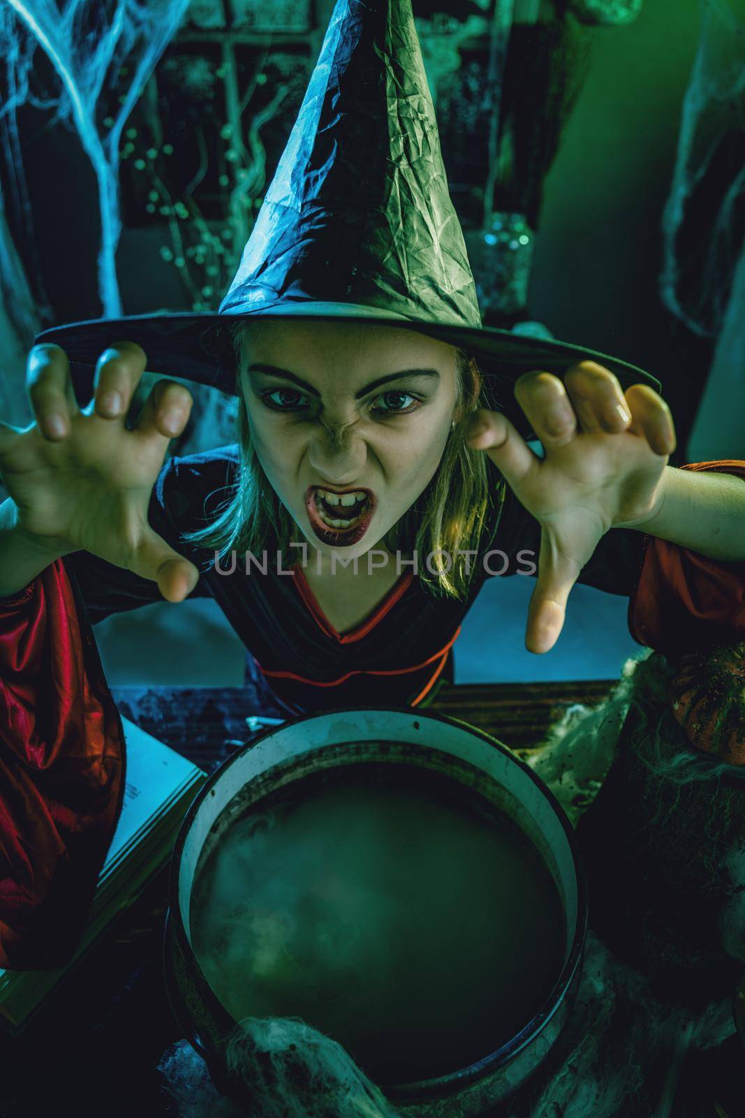 Top view of young witch with awfully face looking at camera and tells evil words above cauldron.