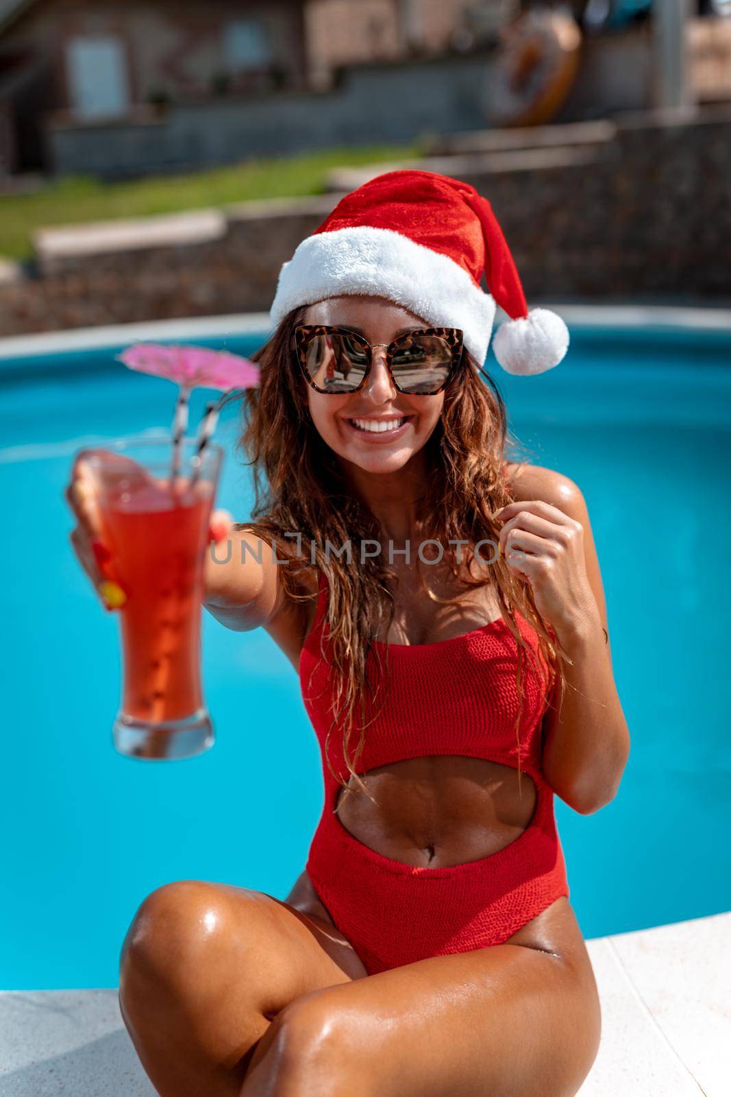 Young beautiful woman near the swimming pool in Santa Claus hat celebrating New Year and Christmas in hot country with glass of cocktail.