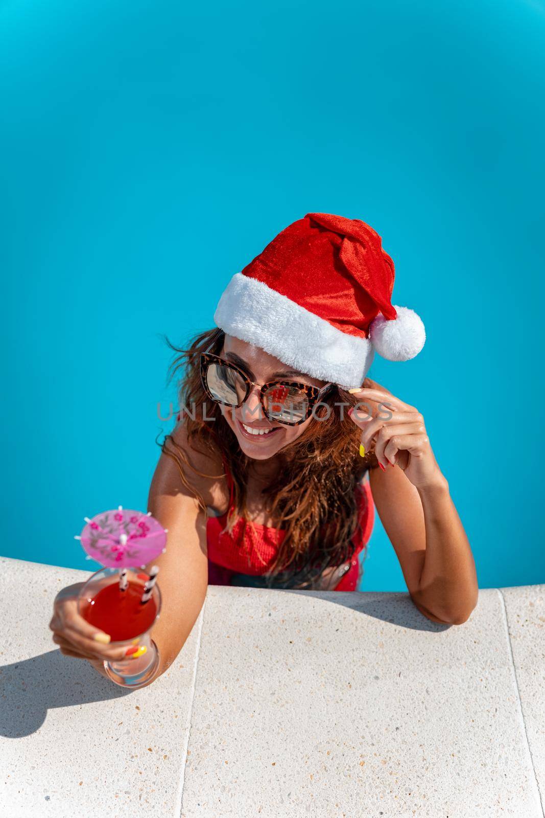 Young beautiful woman in the swimming pool in Santa Claus hat celebrating New Year and Christmas in hot country with glass of cocktail.