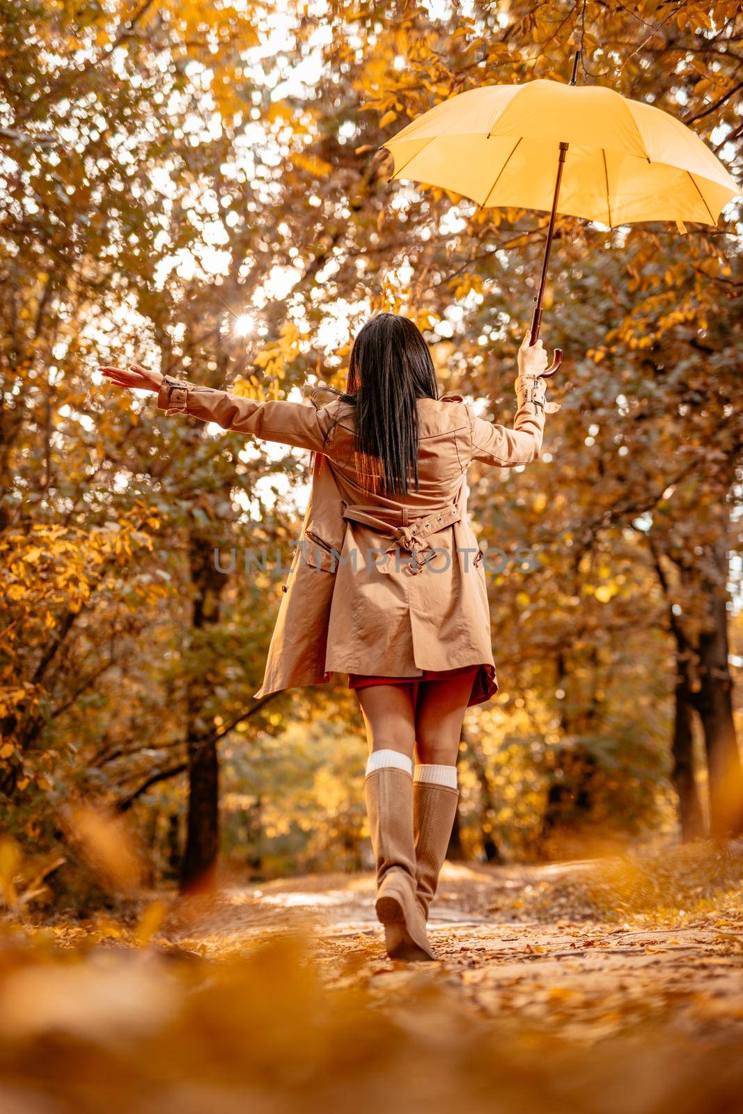 Cheerful young beautiful woman going alone with yellow umbrella in the park in golden autumn. Rear view.