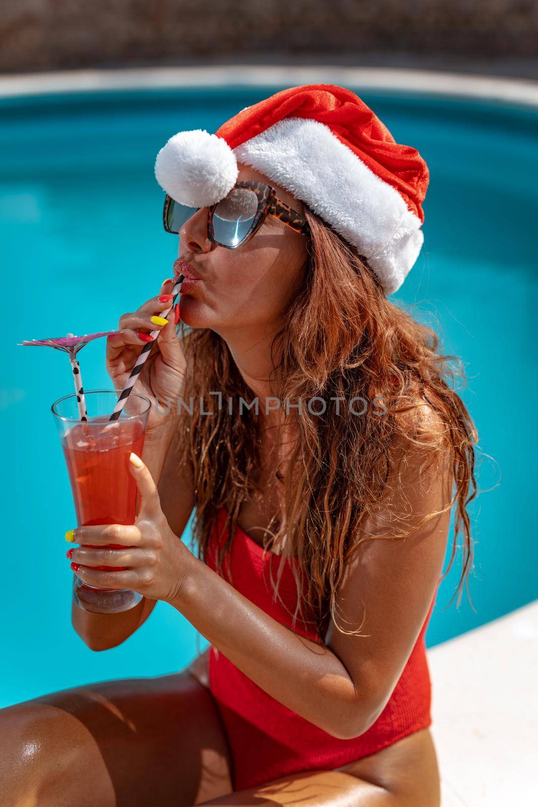 Young beautiful woman near the swimming pool in Santa Claus hat celebrating New Year and Christmas in hot country with glass of cocktail.
