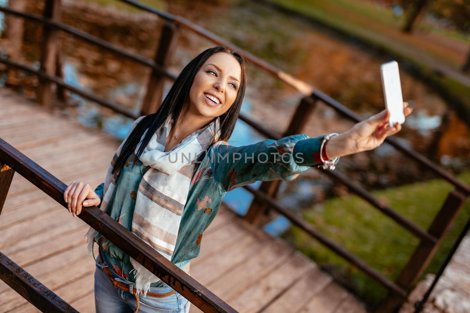 Modern cute girl enjoying to take a selfie on the bridge in autumn city park.  