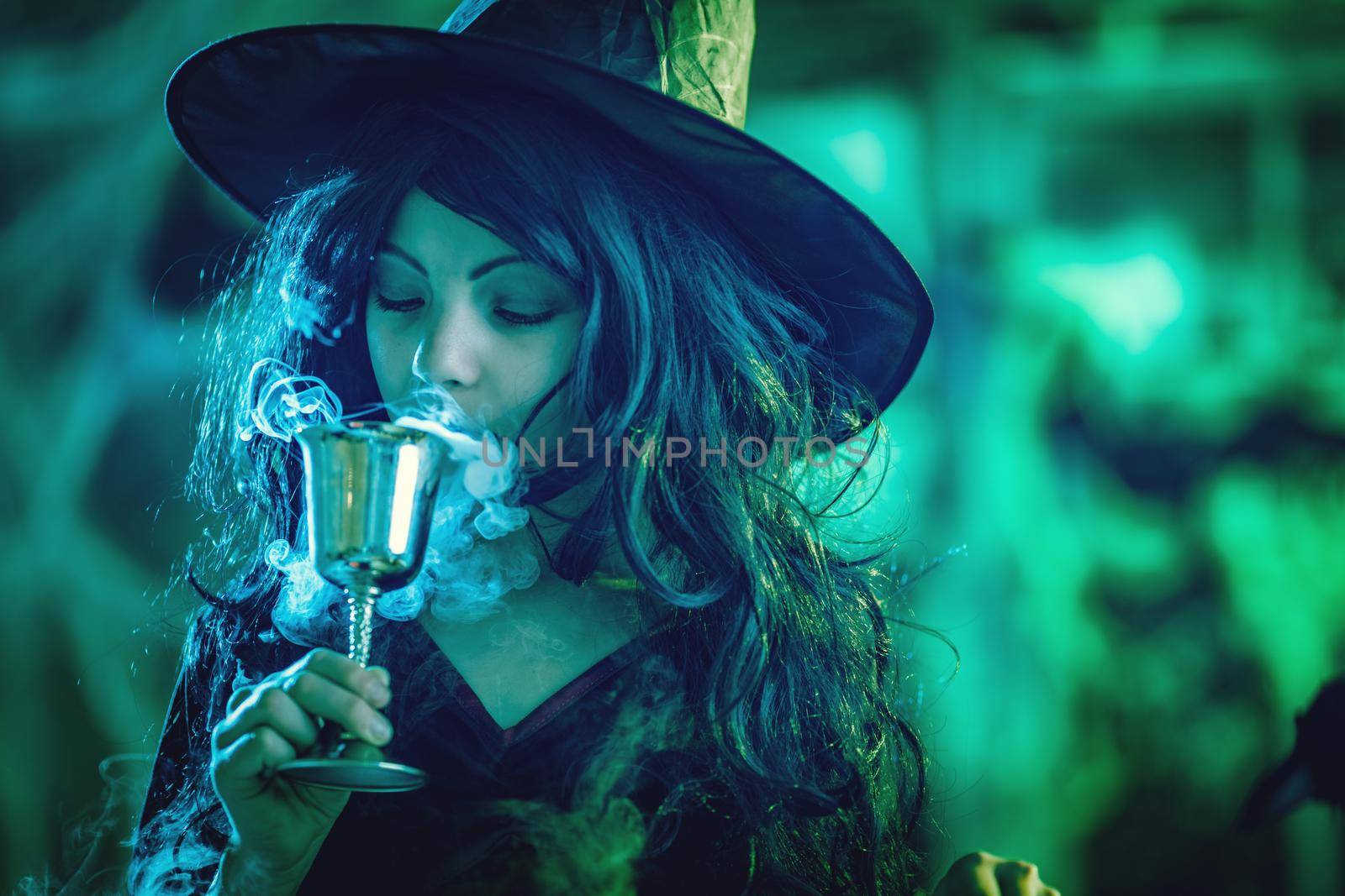 Young witch with serious face holding a goblet and drinks a magic potion in creepy surroundings and smoky green background.