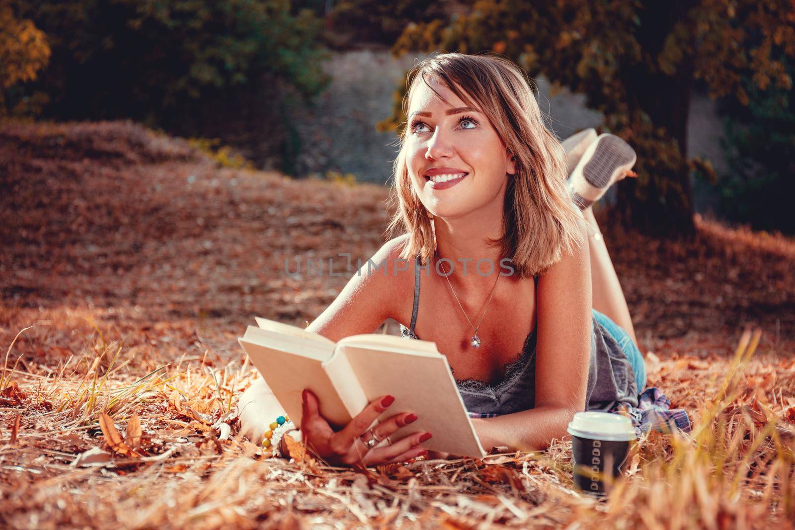 Cute smiling young woman lying down on the withered grass in early autumn sunny day with book.