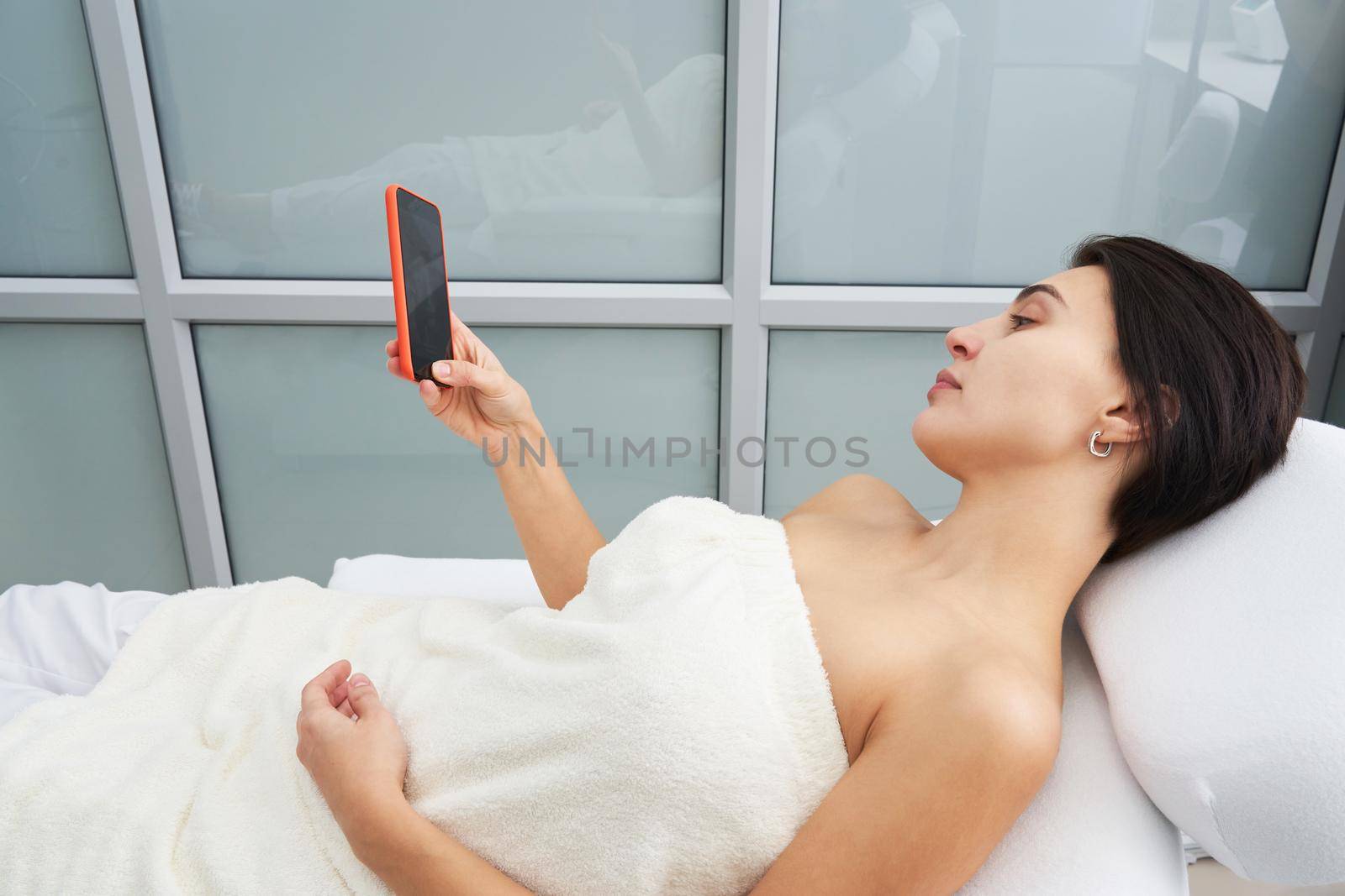 Attractive young woman using a smartphone and texting while laying on a massage bed at spa