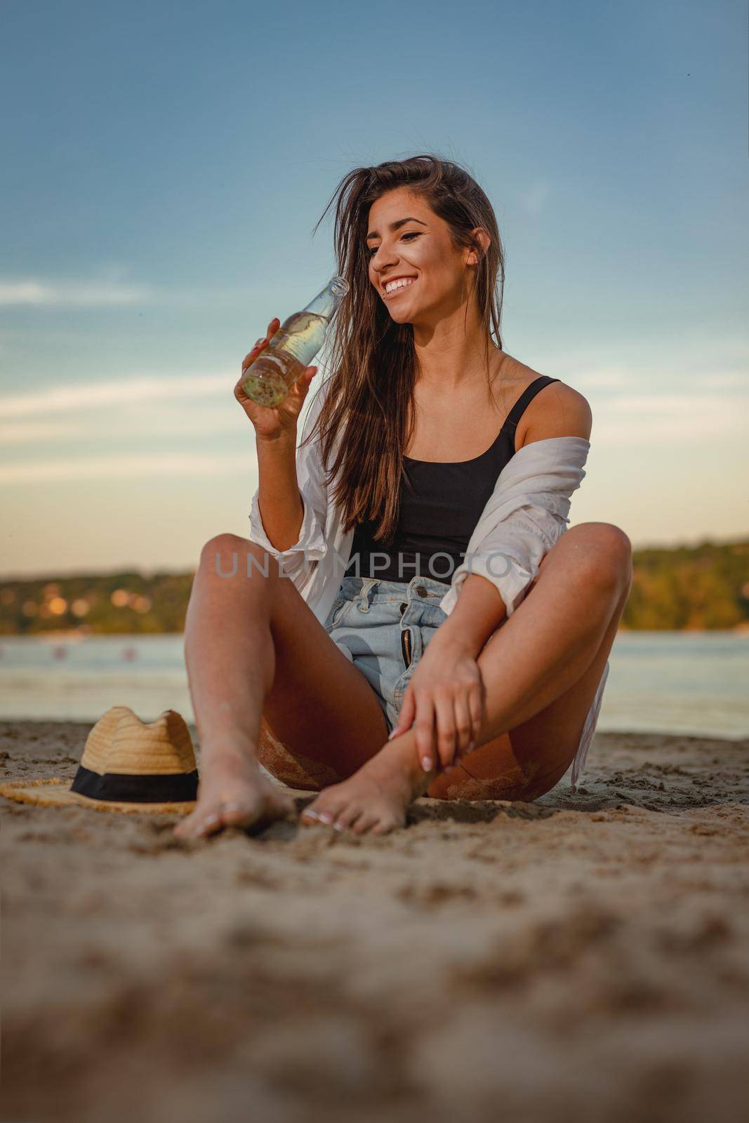 Young woman drinking beer and relaxing at sunset time on the river bank. She is sitting by the river and having fun. 