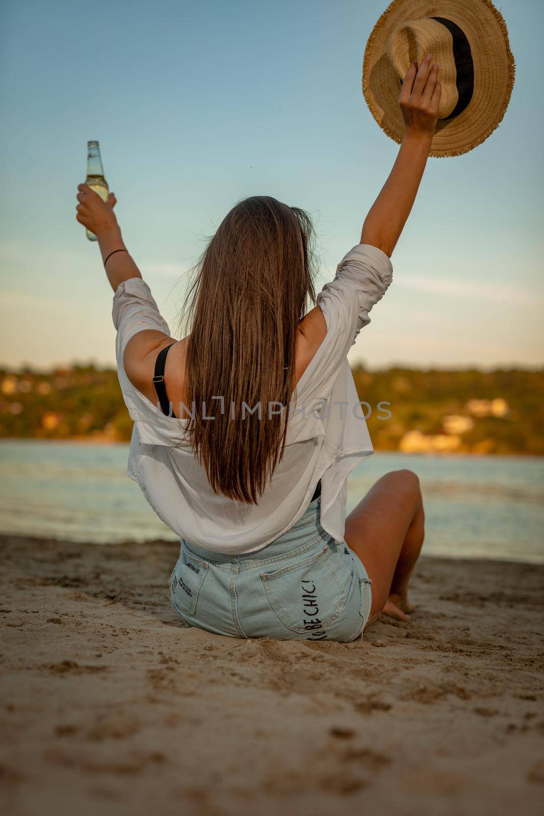 Rear view on a young woman with bottle of beer and straw hat relaxing at sunset time on the river bank.