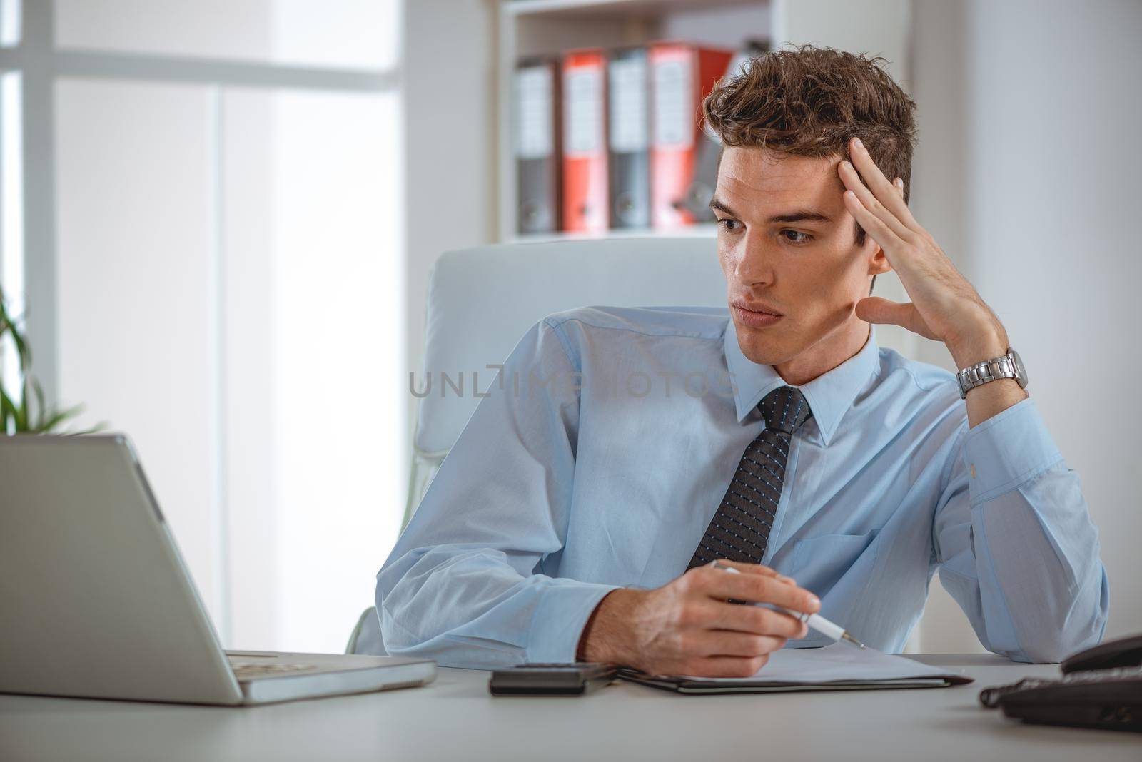 A serious young employee working in office with laptop and making notes, looking focused and concentrated.