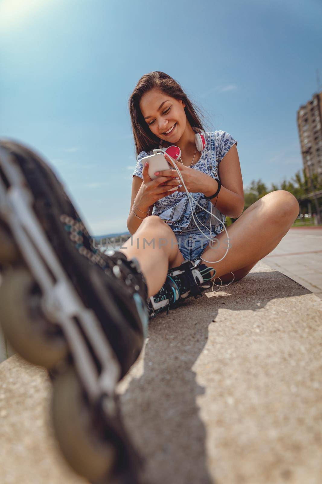 Cute smiling young Filipino woman surfing on internet or typing message on smartphone. She is sitting with roller skates and enjoying on city riverbank at the beautiful summer day. 