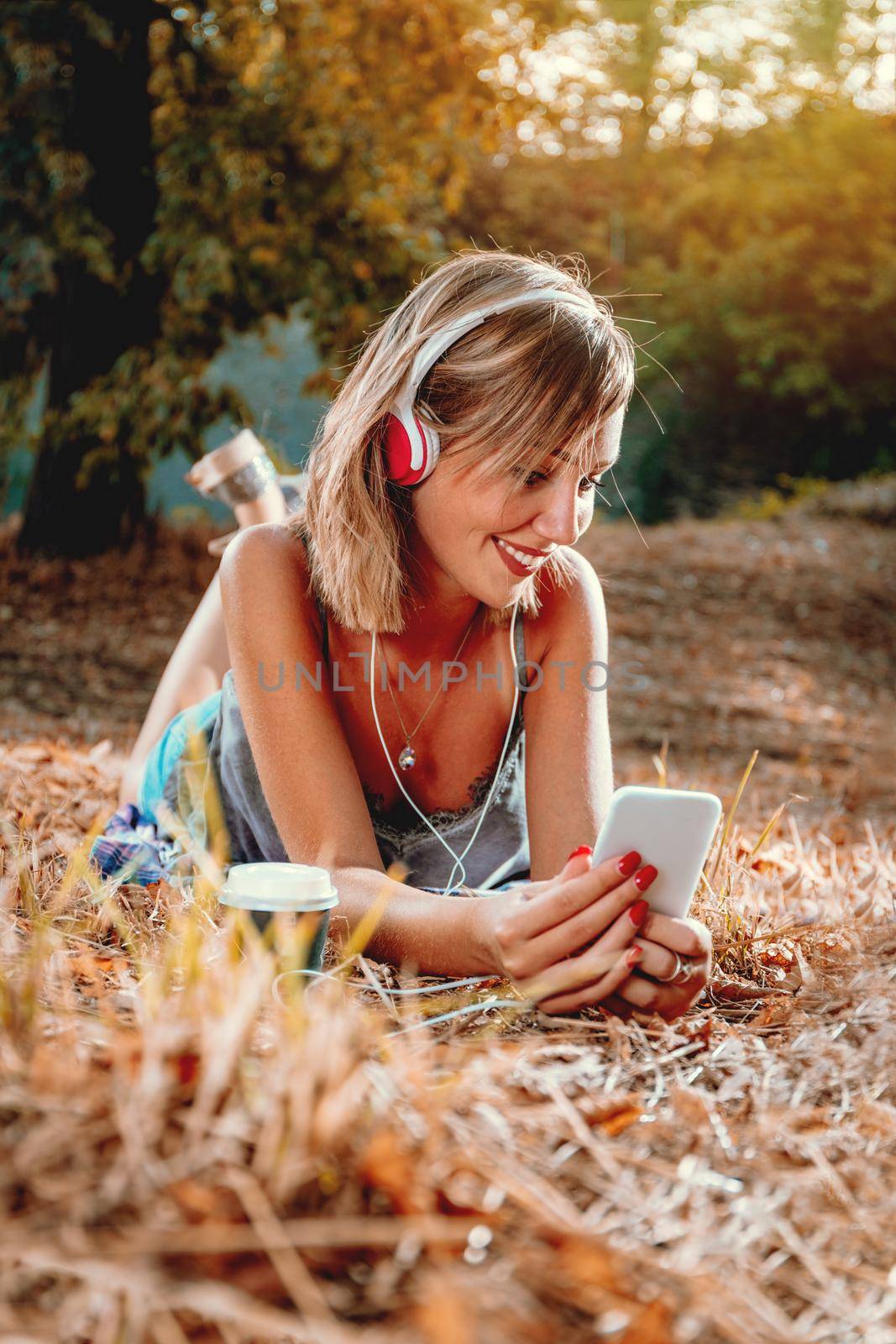 Cute smiling young woman lying down on the withered grass in early autumn sunny day, listening music.
