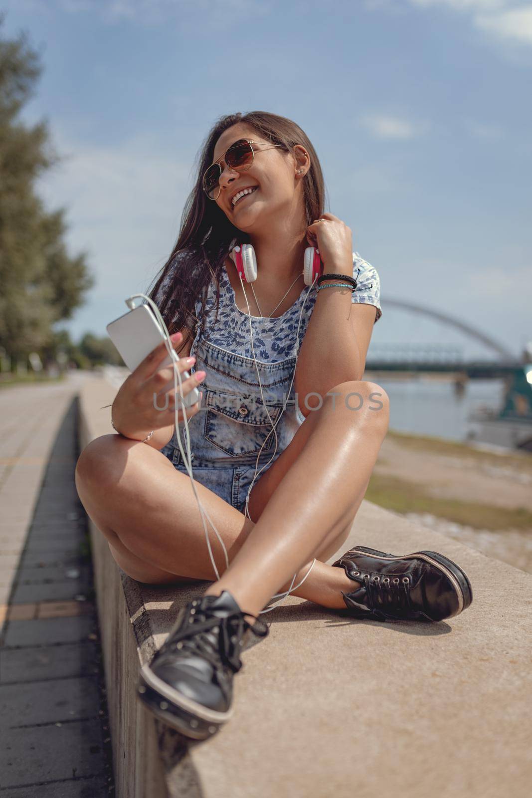A beautiful smiling young woman having fun in the open and enjoying on the city rivershore on a beautiful summer day. She is sitting and holding smartphone and headphones. 