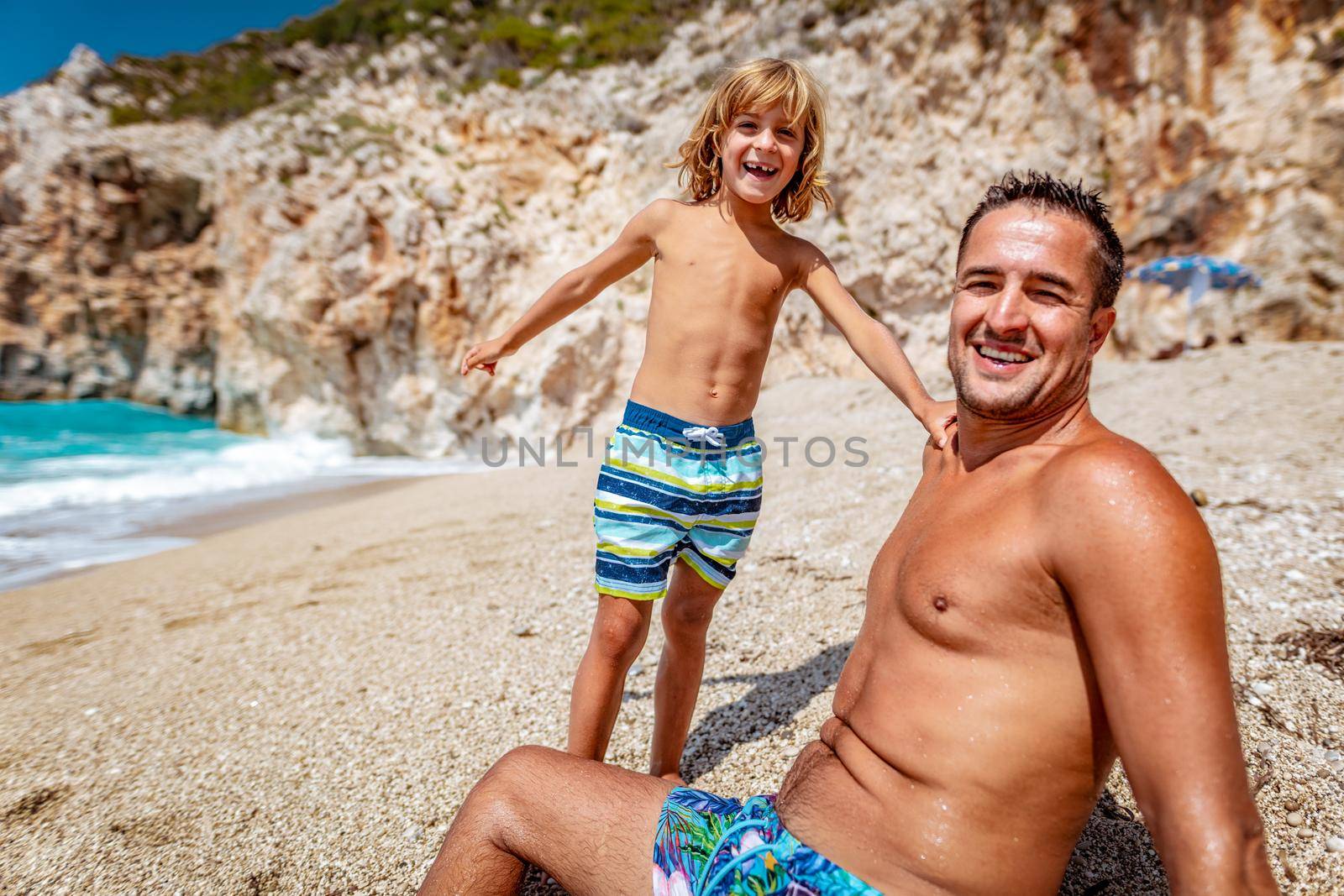 Beautiful little boy enjoying with his father on the beach. They are sitting on the coast and looking at camera.