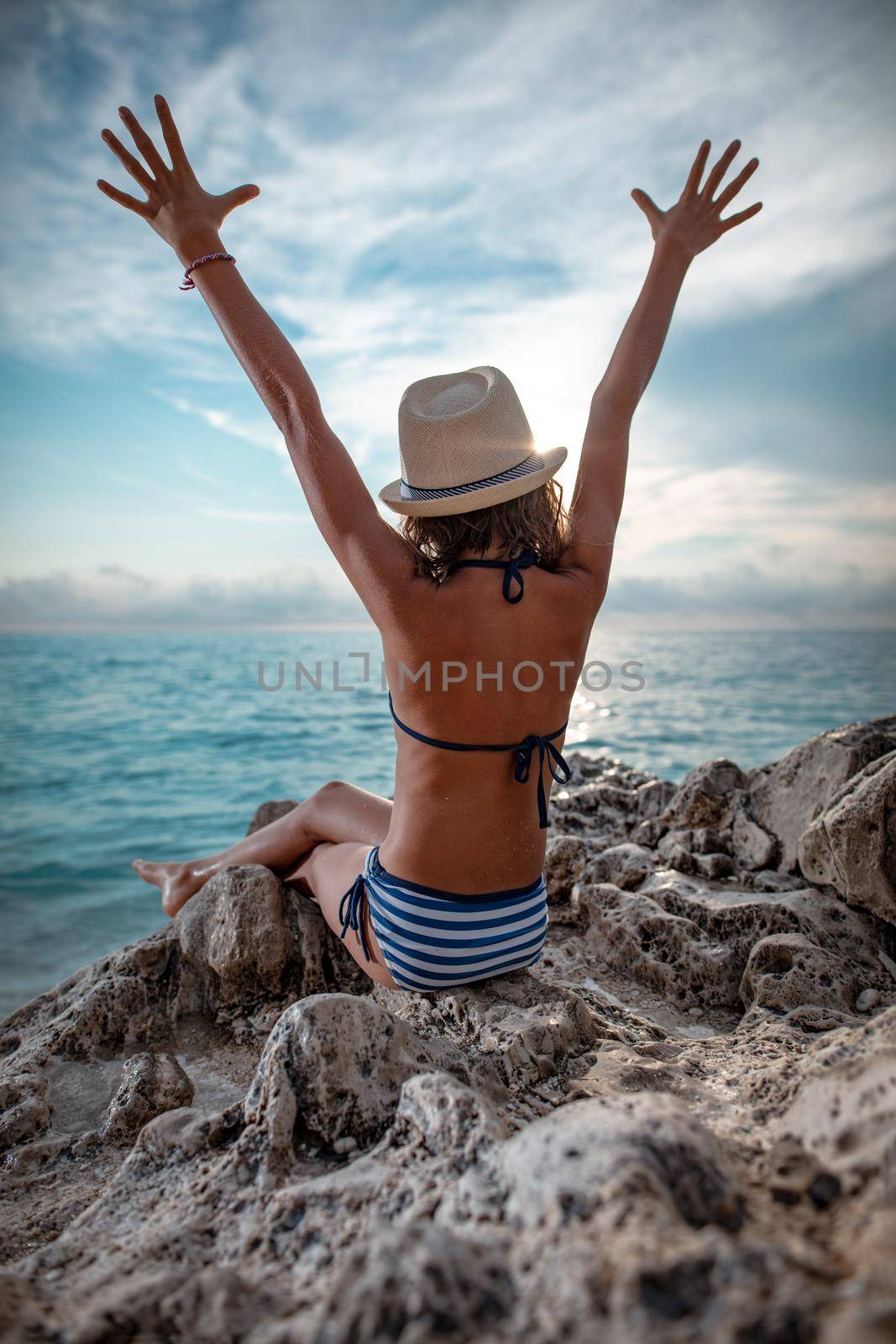 Cute little girl is sitting on the sea rock on the beach. Her arms are high open.  
