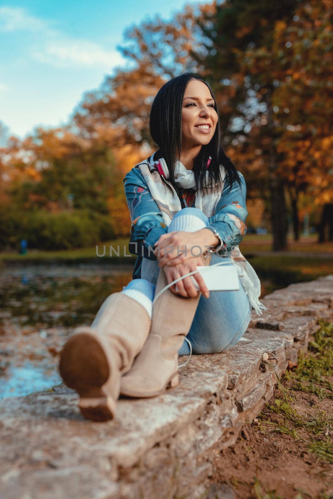 Young woman with smarthphone in her hand and headphones over her neck sitting in a park in a wonderful autumn day.