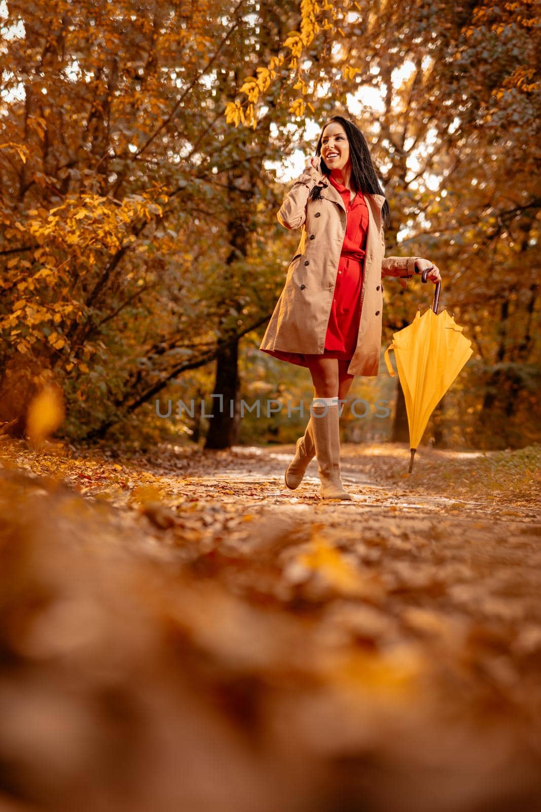 Happy young woman in red dress talking on smartphone walking in autumn sunny park, holding folded yellow umbrella.