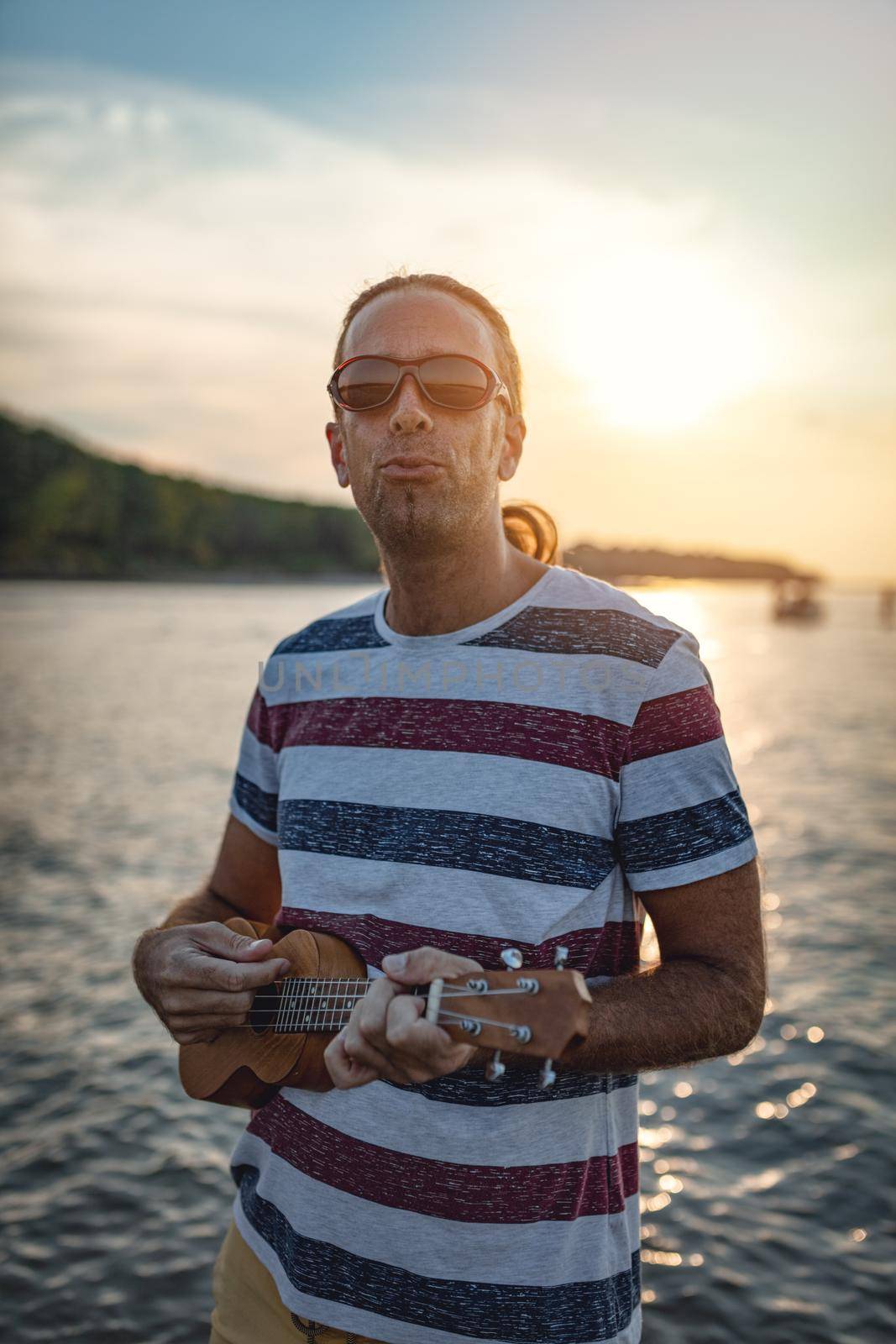 Happy young musician has a great time at the beach. He is standing by the water, playing ukulele and singing. Sunset over water.