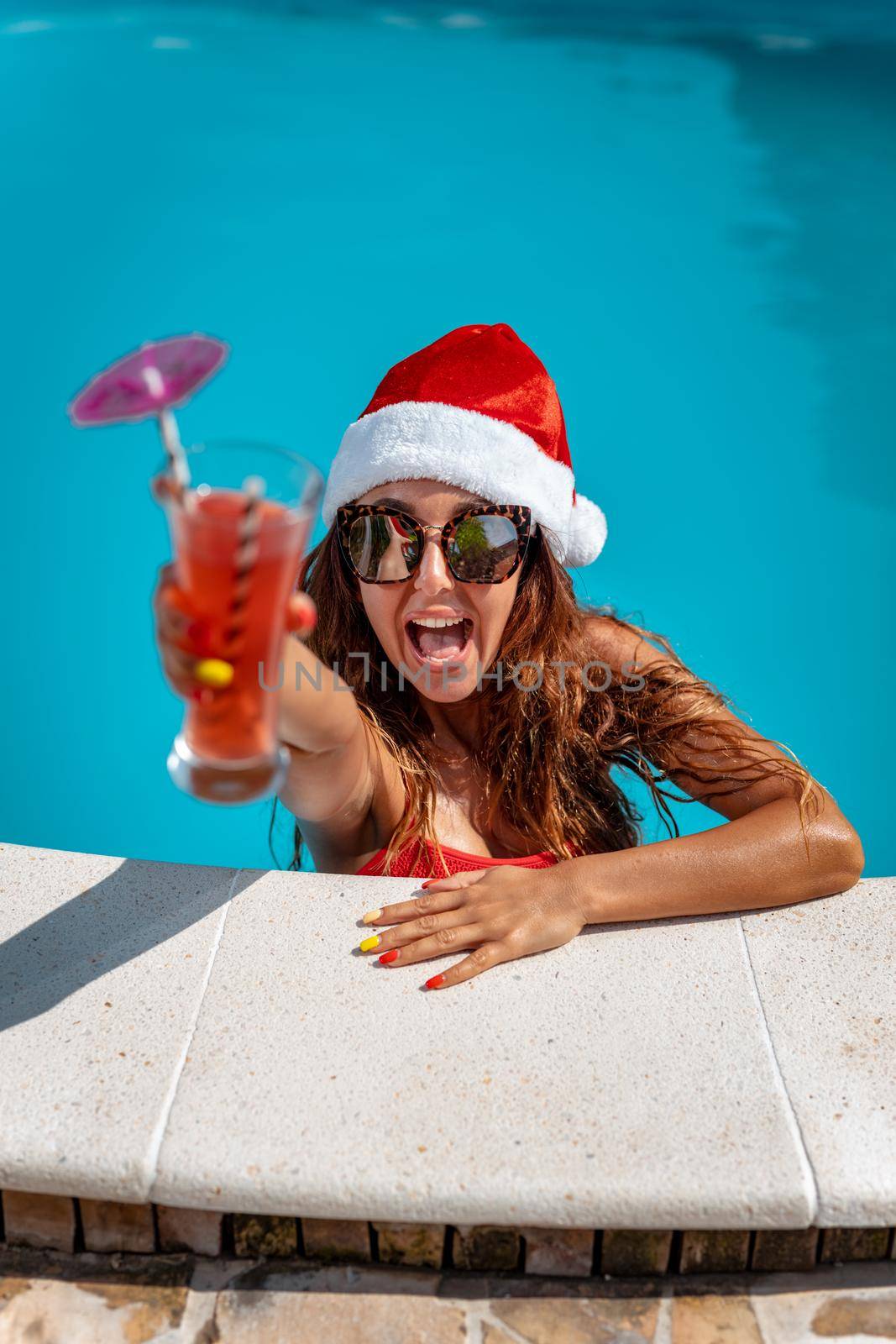 Young beautiful woman in the swimming pool in Santa Claus hat celebrating New Year and Christmas in hot country with glass of cocktail.