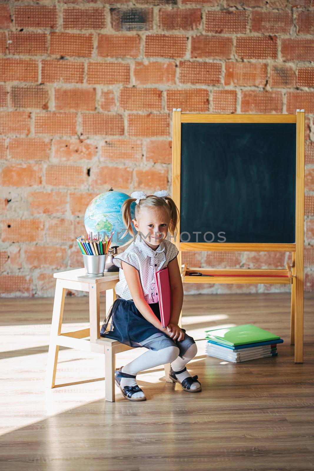 Cute little girl in school uniform sitting next to blackboard with a book, back to school concept