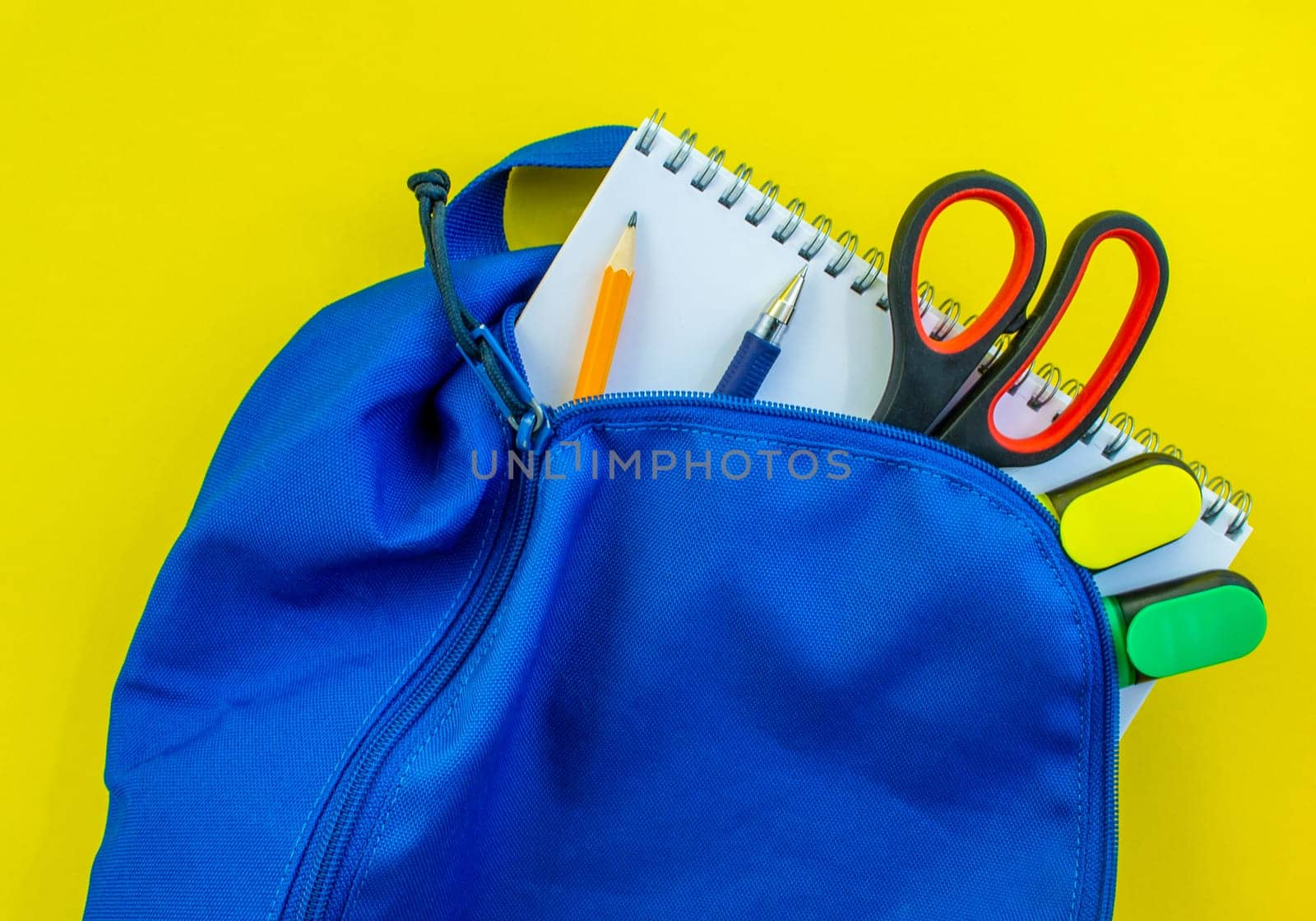 School backpack, pencil, pen, markers, scissors and notepad on a yellow background. School blue backpack with notepad, pencil, markers, scissors and pen inside on a yellow background.