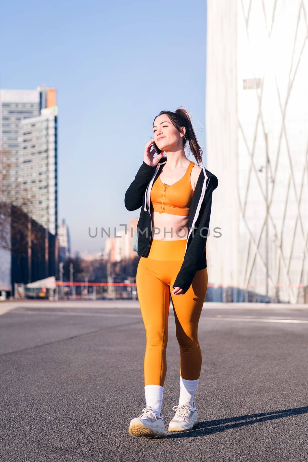 vertical photo of a woman in sports clothes talking by mobile phone outdoors in the city street, concept of technology of communication and urban sport