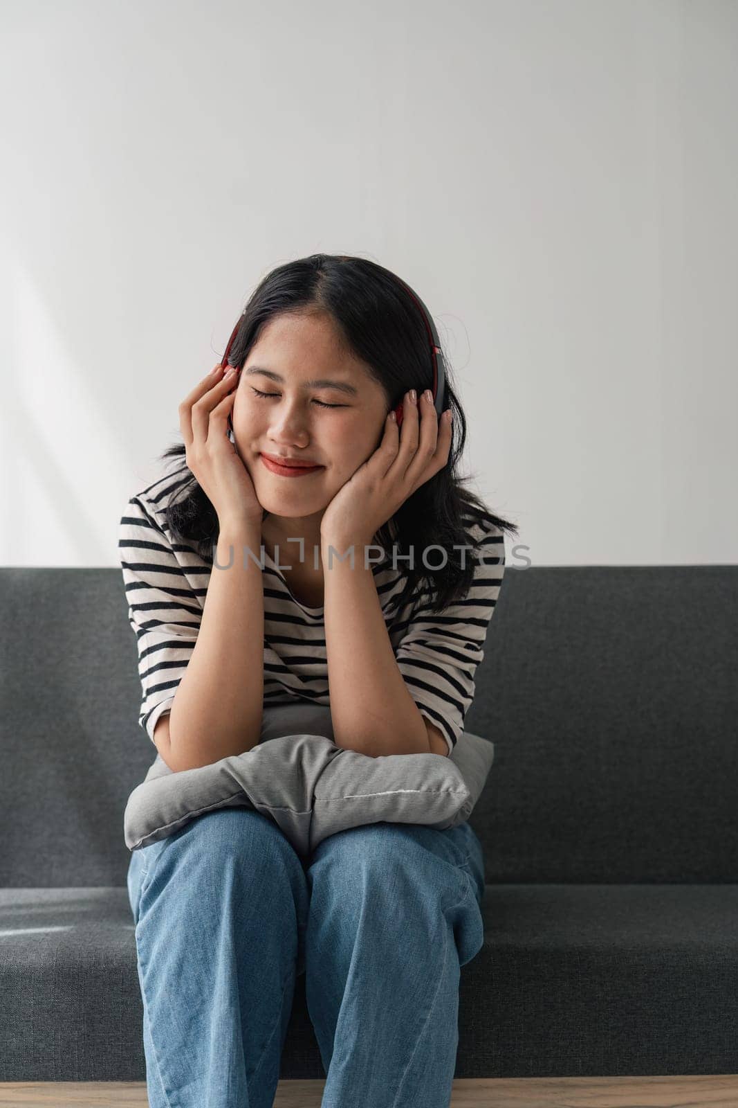 Attractive happy young Asian woman listening music with headphones from music application on laptop and telephone, sitting on sofa by nateemee