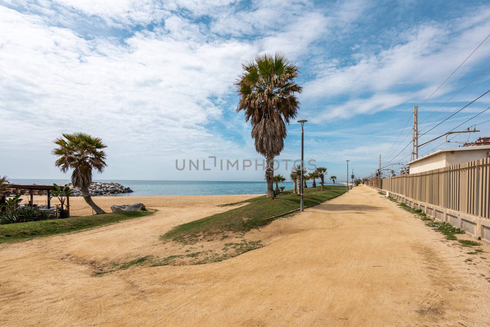 A serene coastal scene in Vilassar de Mar, Spain, with sandy beach, palm trees, and sparkling sea under a clear blue sky.