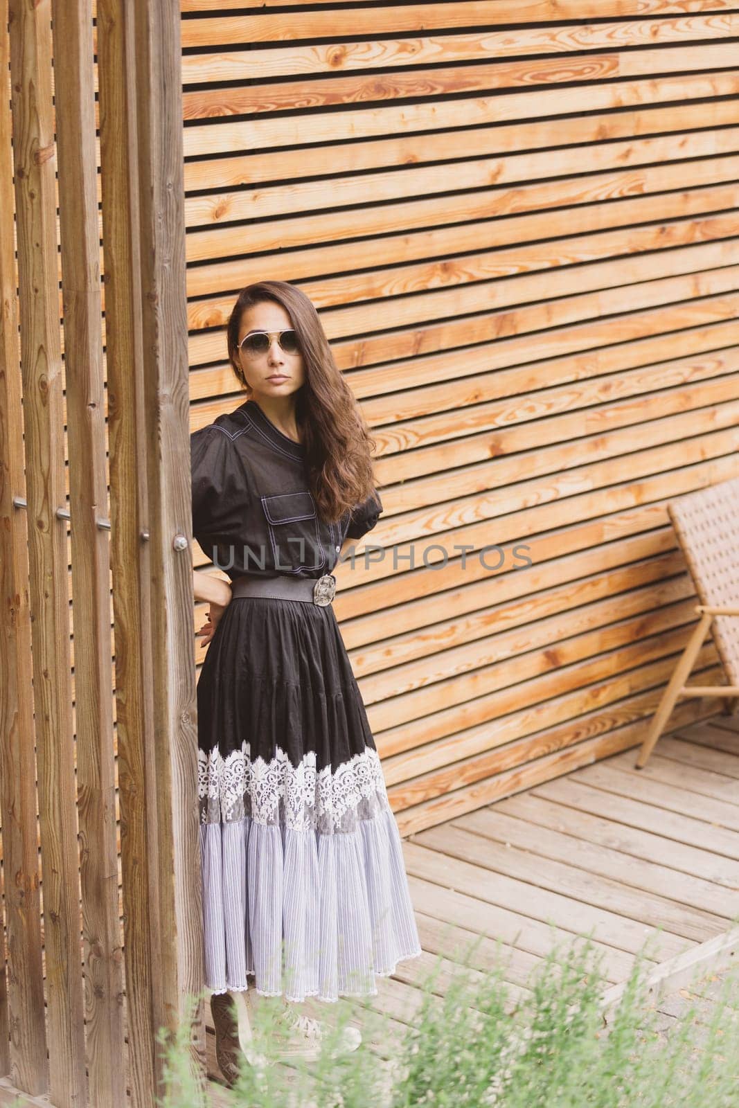 Lifestyle fashion portrait of young stylish hipster woman standing on the street, wearing trendy outfit, black dress. Wood background. by sarymsakov