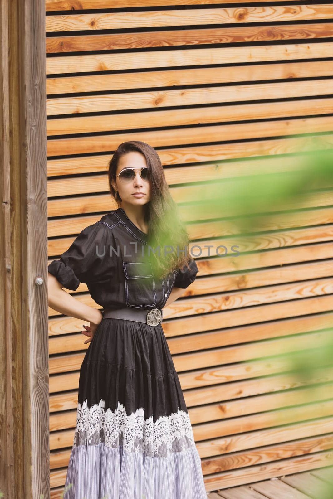 Lifestyle fashion portrait of young stylish hipster woman standing on the street, wearing trendy outfit, black dress. Wood background. by sarymsakov
