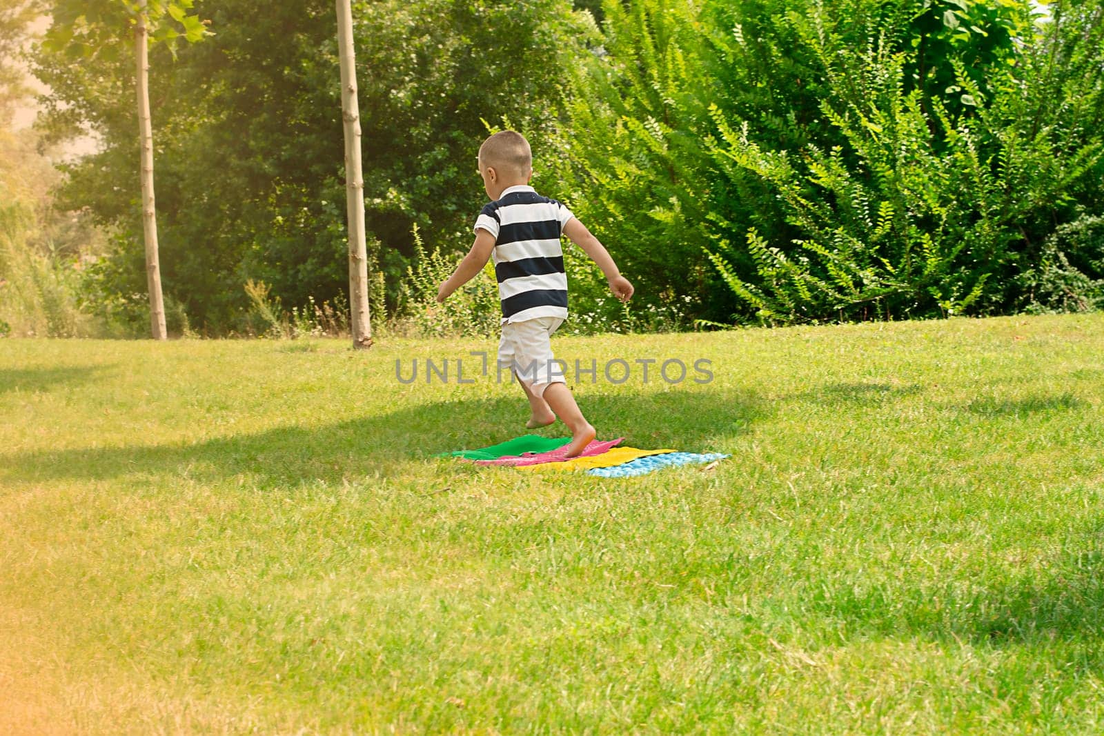 A child cheerfully runs barefoot on massage, multi-colored orthopedic rugs in the fresh air, in a green park in summer. Health concept. copy space.