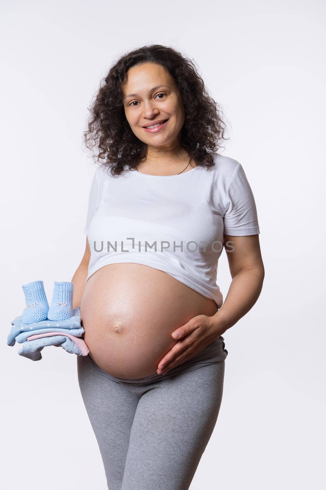 Adult pregnant woman smiles looking at camera, stroking her big belly, holding pile of baby clothes on white background by artgf