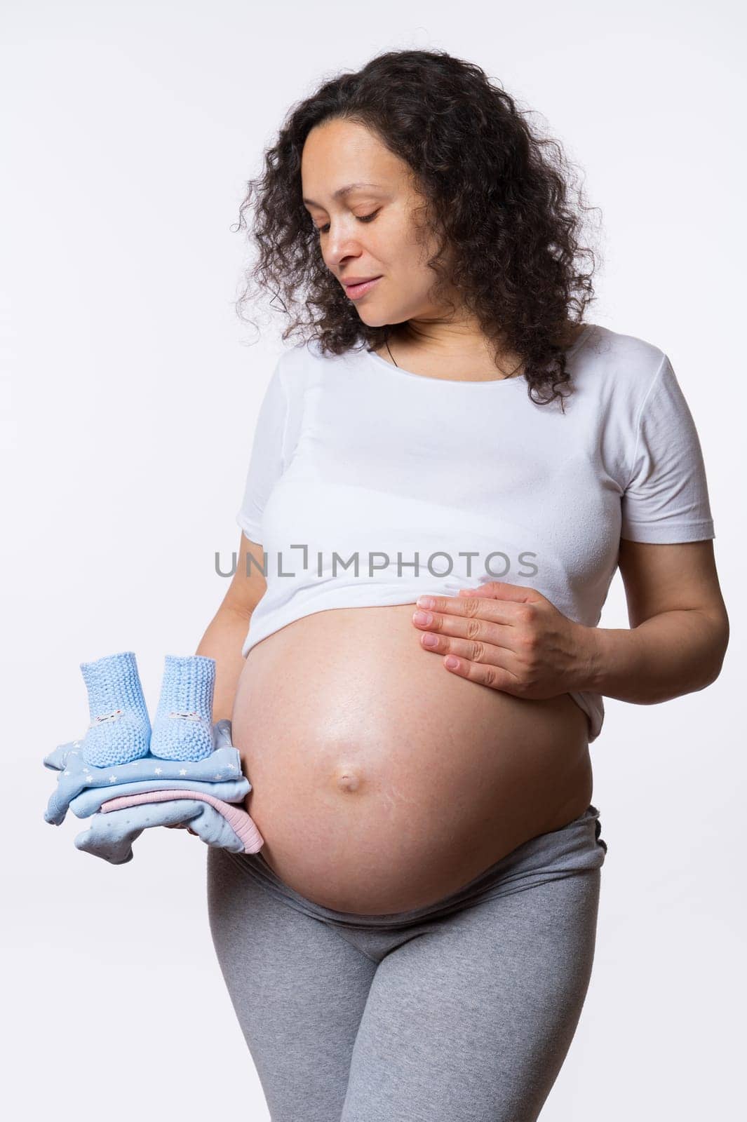 Beautiful expectant gravid mama, multi ethnic adult happy pregnant woman holding newborn clothes, posing with bare belly, isolated on white studio background. Pregnancy. Maternity. Expecting a baby