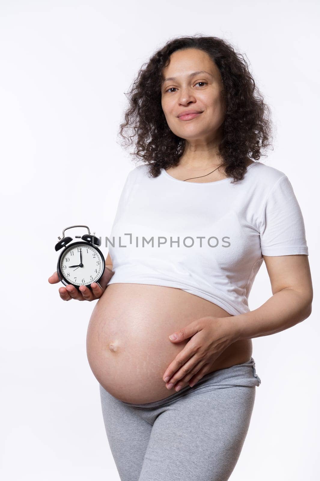 Charming pregnant woman gently strokes her belly, cutely smiles, shows alarm clock to camera with ticking biological time and countdown to the expected date of child birth. Waiting for baby. Pregnancy