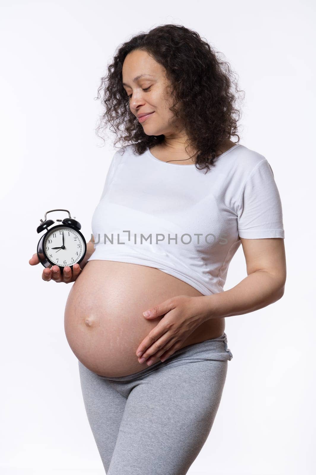 Happy delightful curly haired beautiful pregnant woman, expectant gravid mother caressing her tummy belly, holding alarm clock, isolated white studio background. Pregnancy and Baby's due date concept