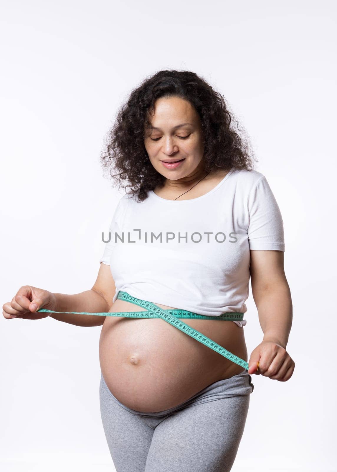 Advertising studio portrait of a curly haired adult pregnant woman, expectant mother, monitoring the development of her baby in womb, measuring her big belly with a tape, isolated on white background